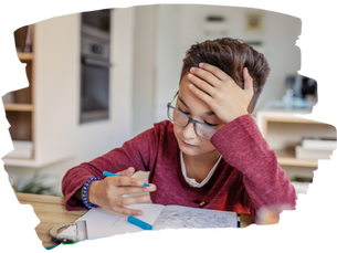 Boy in red sweater looks frustrated while doing homework at a wooden table. He holds a pen and rests his head on his hand.