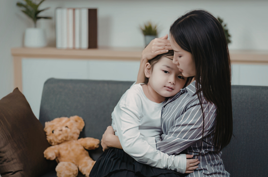 Woman holds and kisses a sad child on a gray sofa in a cozy room. A teddy bear is nearby. Warm, comforting atmosphere.