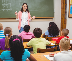 Teacher in pink cardigan instructs attentive students in a classroom. Chalkboard with math problems in the background. Bright, engaged setting.