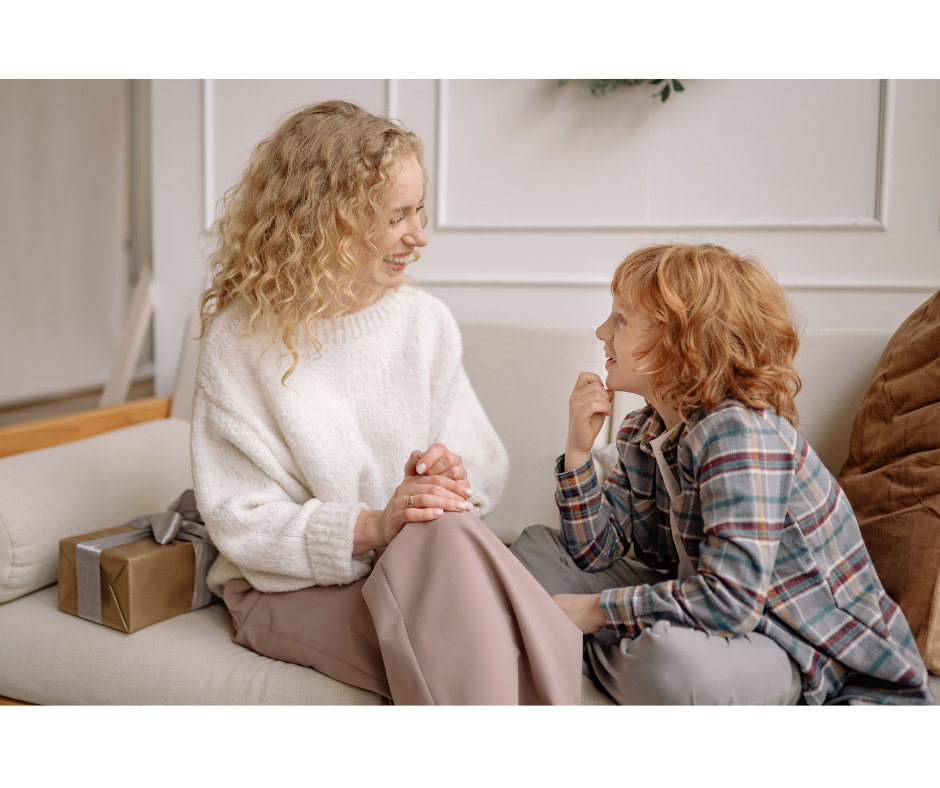 Woman and child smile at each other on a cozy sofa. Gift box beside them. Light, warm room with white walls. Joyful mood.