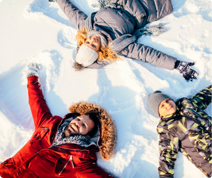 Three people making snow angels, lying on their backs in colorful winter gear. The scene is bright and cheerful in a snowy landscape.