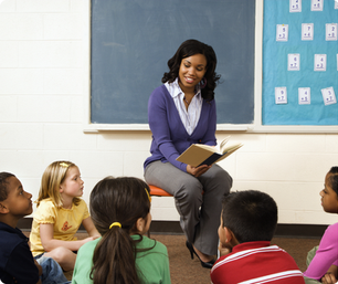 Teacher reads to attentive children in a classroom. Background shows math cards on a blue board. Teacher wears purple, children sit on carpet.