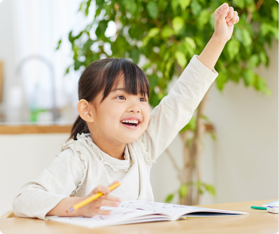 Smiling child raises hand enthusiastically while sitting at a desk with an open book and pencil. Bright room with green plant in background.