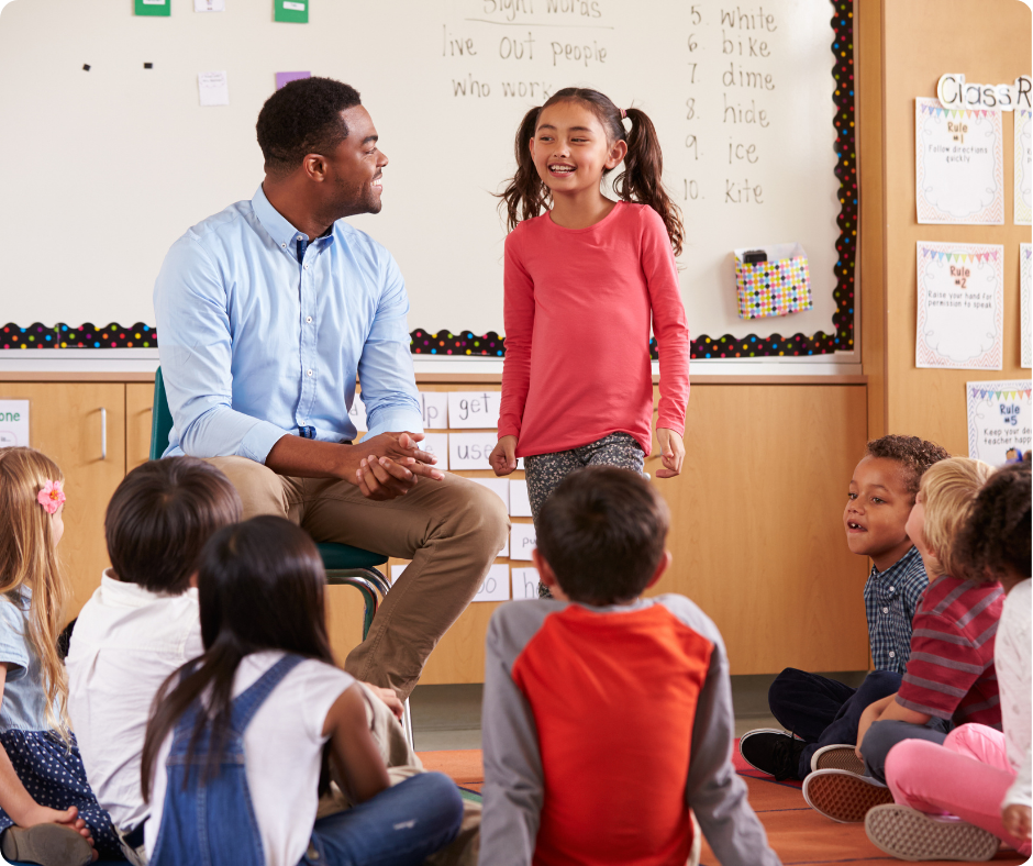 Teacher and students in a classroom. A girl stands smiling, surrounded by seated children. A whiteboard with writing is in the background.