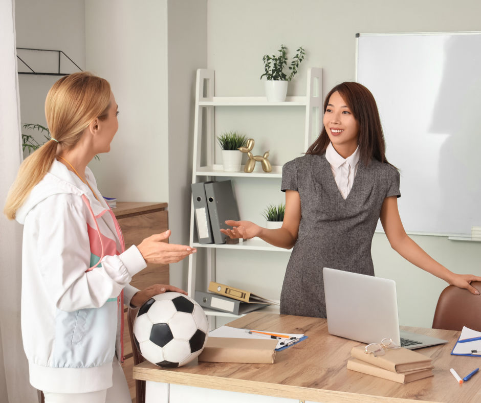 Two women in an office chat joyfully. One holds a soccer ball. A laptop, books, and pens are on the desk. Whiteboard and shelves in back.