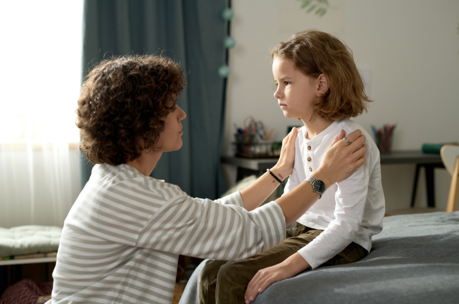 Adult and child sit on a bed in a cozy room. The adult gently holds the child's shoulders, creating a calm, comforting atmosphere.