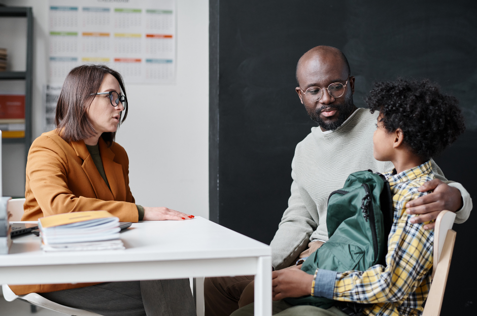 Counselor speaks with a father and child in an office, the father hugging the child with a backpack, calm and serious.