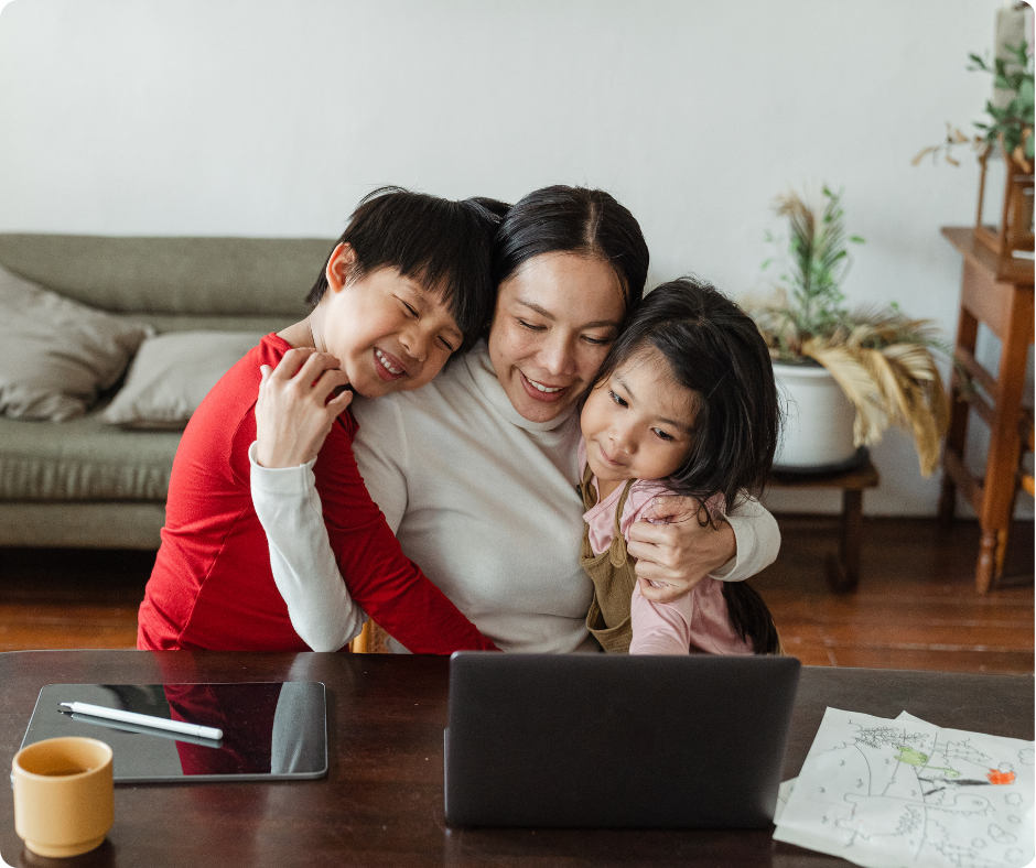 Woman and two children hugging and smiling at a laptop on a table. Warm home setting with a sofa, plants, and papers. Cozy and joyful mood.