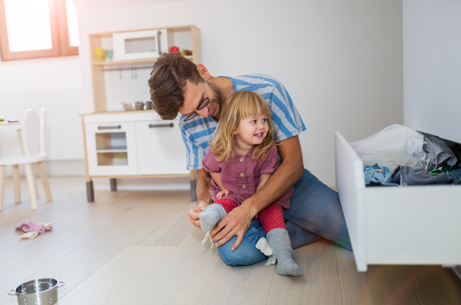 Father hugs smiling young girl on floor beside open laundry drawer in a bright kitchen/playroom.