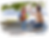 Child with backpack high-fives woman outdoors. Child holds books, wears red shirt. Both smiling under a leafy tree. Casual scene.