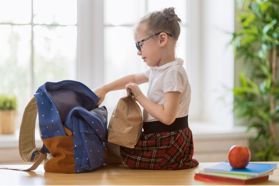 Girl with glasses packs lunch in a brown bag into a blue backpack. She's indoors, wearing a plaid skirt. Apple and notebooks on table.