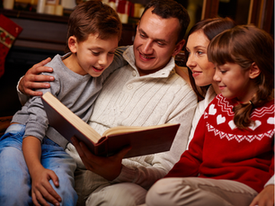 Family of four reading a book together on a couch. Cozy setting with warm red and white clothing, evoking a joyful, festive mood.