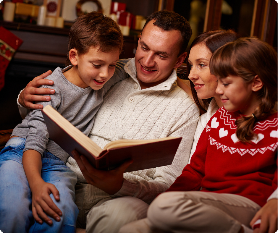 Family of four reading a book together on a couch. Cozy setting with warm red and white clothing, evoking a joyful, festive mood.