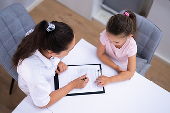 Woman and young girl review paperwork at a white table, the woman writes on a clipboard, indoors with gray chairs.
