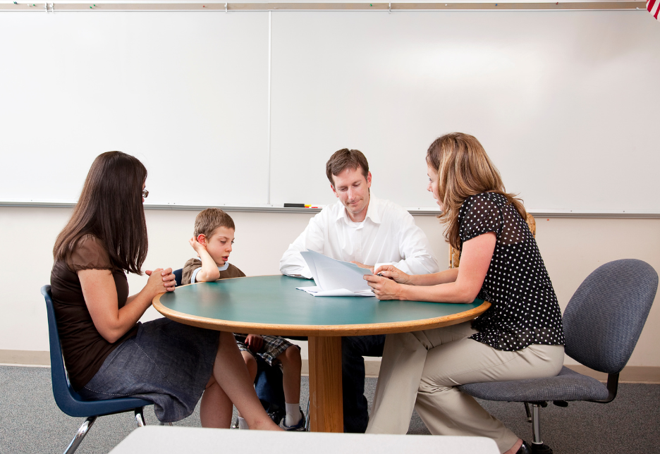 Teacher and parents meet with a young boy at a round table in a classroom, reviewing papers in front of a whiteboard.