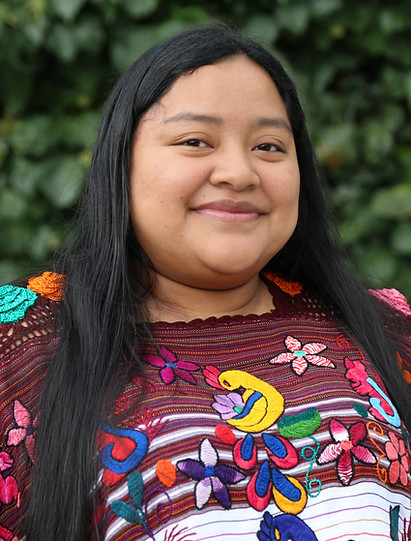 Smiling warmly before a lush backdrop, Shirly is framed by a tapestry of deep green leaves.
