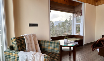 Cozy room sofa, coffee table, pinewood ceiling, large bay window with seating overlooking mountain trees.