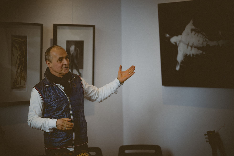 Marek in blauer Weste erklärt Kunstwerke an der Wand in Galerie. Eine Hand zeigt auf das Bild Heldenschmiede, ruhig und konzentriert, gedämpfte Beleuchtung.