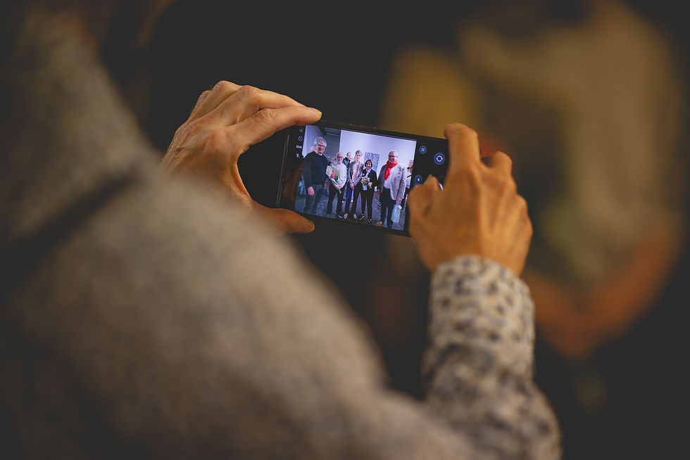 Hand hält Smartphone, fotografiert eine Gruppe lächelnder Menschen vor heller Wand. Person im roten Schal. Stimmung ist freudig.