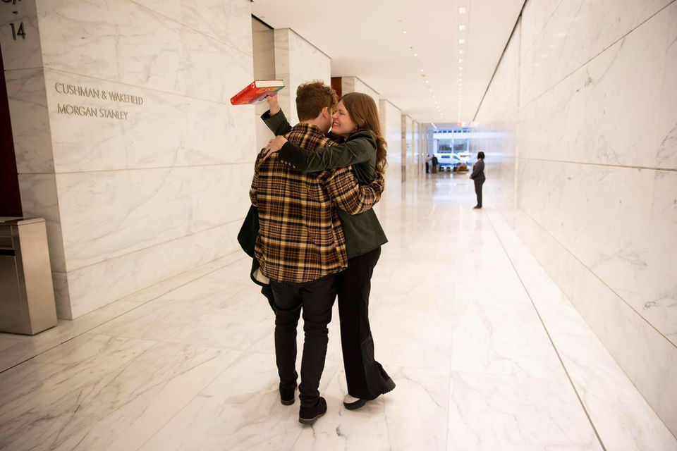 Photo of Sophia hugging her husband in a long hallway with white stone floor and walls. She is grinning ear to ear and he is hugging her back.