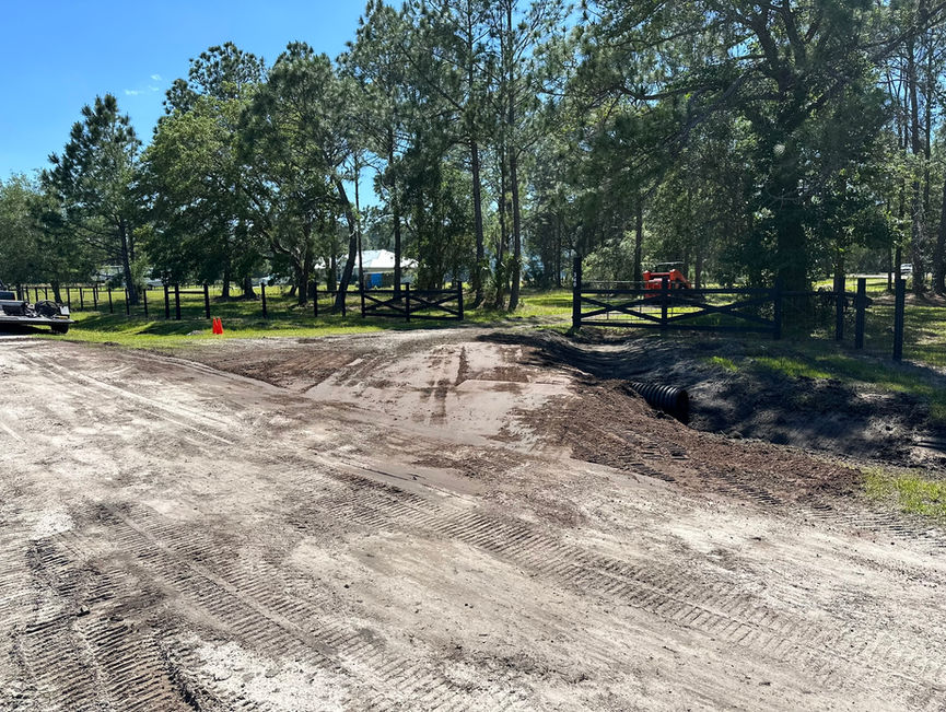 A sandy pine lot with a new culvert pipe, a black farm gate, and a tracked machine parked among the pines in New Smyrna Beach, Florida.