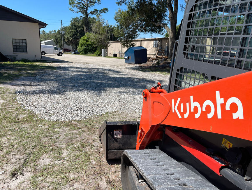 A red Kubota track loader beside a freshly spread gravel pad at a work yard in New Smyrna Beach, Florida.