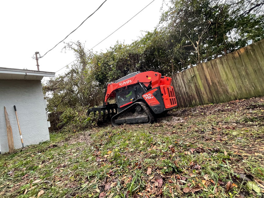 A red Kubota track loader with a grapple clearing brush along a wooden fence beside a house in Edgewater, Florida.