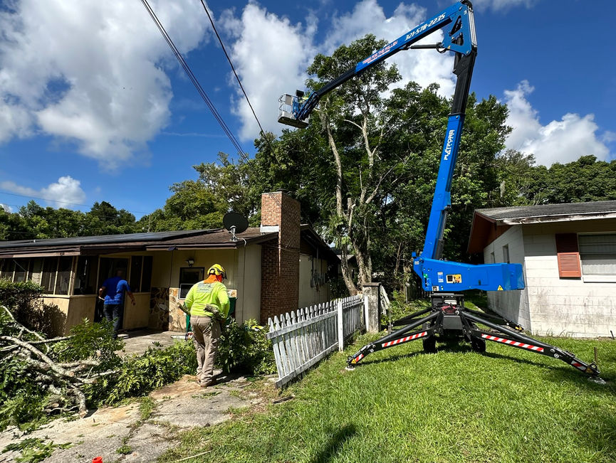 A blue boom lift set on its outriggers in a front yard, with a crew member in green nearby in New Smyrna Beach, Florida.