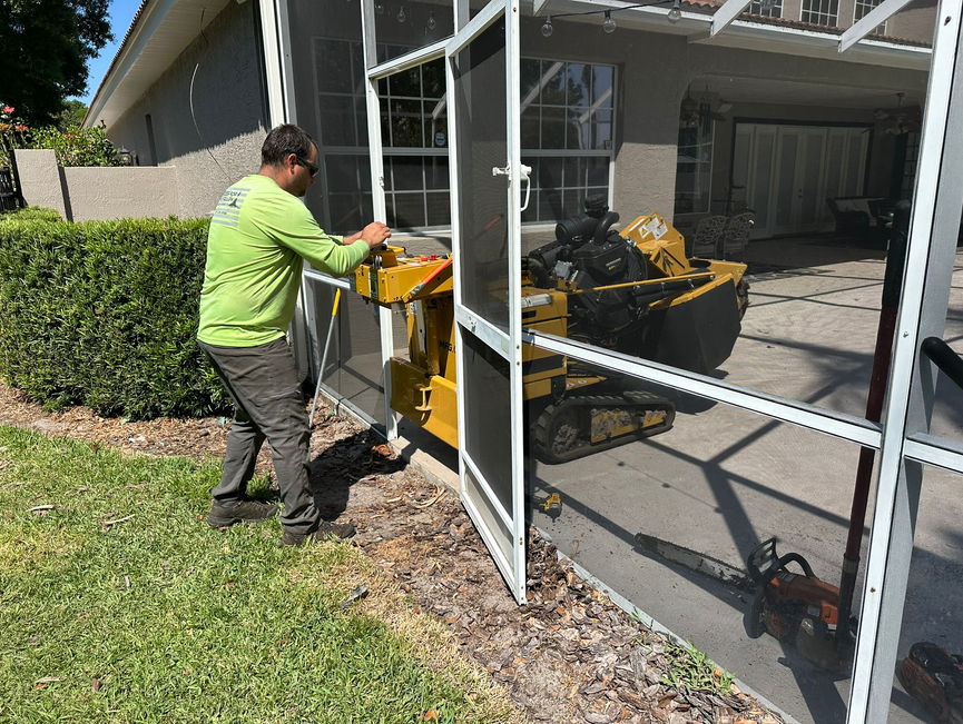 A crew member in a lime shirt pushing a yellow stump grinder through a doorway in Port Orange, Florida.