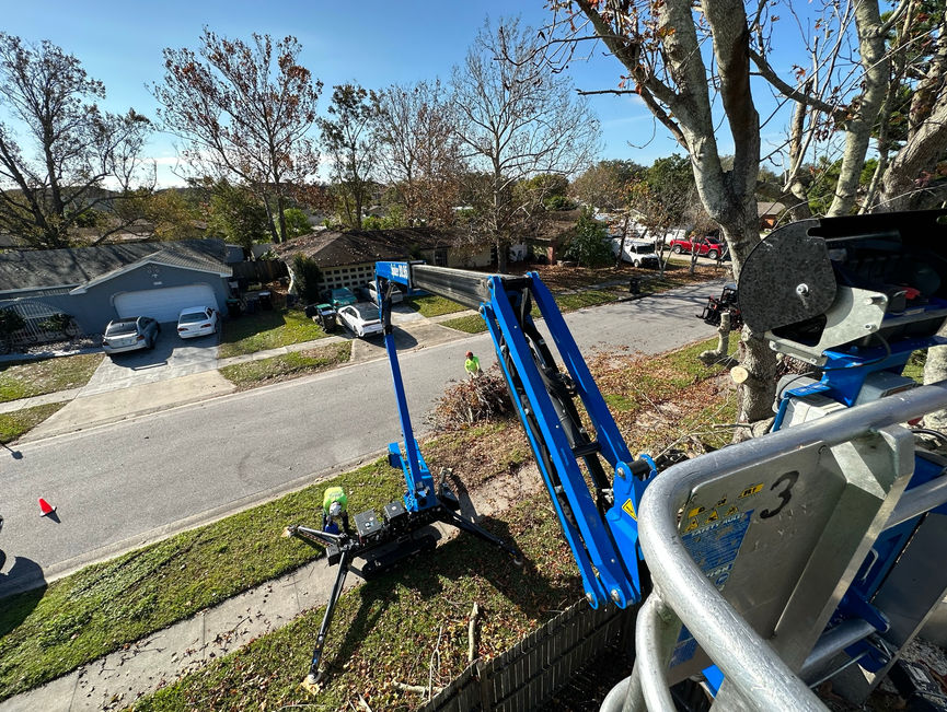 A blue boom lift strapped on a trailer at the curb, with the work truck in the foreground in Port Orange, Florida.