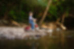 A 18 month old boy sits in a beautiful creek setting with trees behind him and water in front of him. He sits upon a red tricycle looking at the camera while a handmade stick fishing pole is sticking out of the ground in front of him. A bobber floats in the water attached with fishing line to the stick.