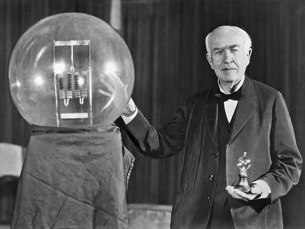 Historic photograph of Thomas Edison seated beside an oversized lightbulb, symbolizing his groundbreaking work in electric lighting.