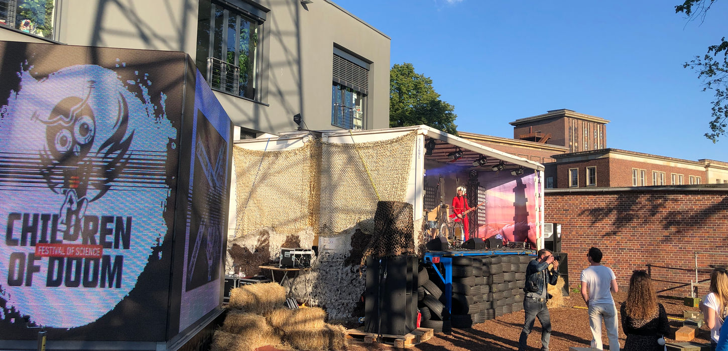 Outdoor-Event-Setup mit 'Children of Doom' Banner und Publikum vor industrieller Gebäudekulisse bei Tageslicht