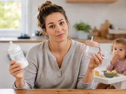 Parent holding a bottle, pacifier, and cup while deciding when to transition a toddler away from bottles and pacifiers