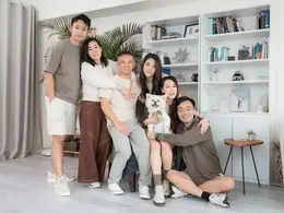 客厅里一家七口与白色小狗合影,围坐在一起微笑,背景有白色书架、绿植和灰色窗帘,氛围温馨。