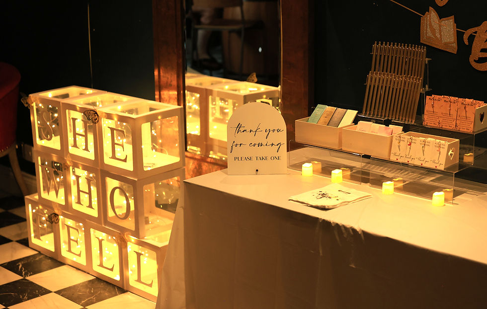 Hayley Stoner's she who fell book launch and live music experience. Ballon boxes for she who fell. There is led lighting in each box and 3d butterflies on the front of some of the boxes. The boxes are stacked, with she on the top, then who in the middle and fell at the bottom. The table has a thank you for coming - please take one sign, two, two tier transparent perspex display boxes with 4 small led candles placed in front of them. On the first tier are 3 wooden boxes that run across both display boxes and contain the complimentary gifts for guests - She who fell branded booked marks, affirmation cards and little heart bag charms that say on the backing card "a little hug to say a big thank you." On the top tier there are gold branded pens saying she who fell and the author website hayleystoner-author.com. The pens are in a pen display stand. Next to them on the other display box is another wooden box with musical note bag charms in.