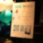 A2 printed board for Hayley Stoner's She who fell book launch and live music experience event captures a photo of Hayley from the front cover of the book, the title a quote "some books are written, some book rewrite you", a QR code to Hayley's website hayleystoner-author.com and a mock up image of the front and back of the book she who fell. The board is on a black easel in the venue next to a balloon stand.