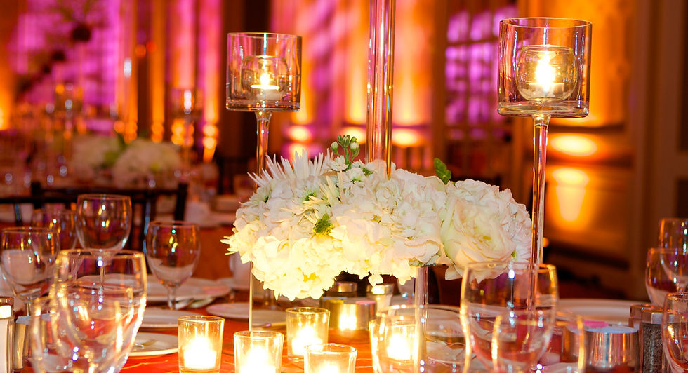 Beautiful venue decoration showing white roses as centrepieces, with tall glass candle holders either side. Stunning warm pink and orange lighting is captured in the background, as well as other tables in the venue.