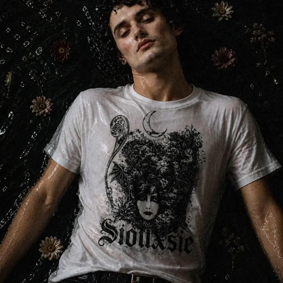 Thumbnail: Menino de camiseta branca com estampa Siouxsie apoiado na parede com papel de parede preto  de flores