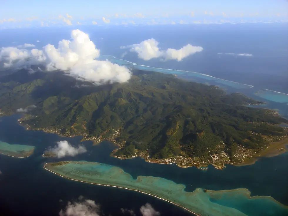Vue aérienne de Raiatea, entourée de lagon turquoise et de récifs, sous de gros nuages blancs.