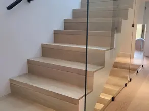 Modern light wood staircase with frameless glass railing and black wall-mounted handrail.
