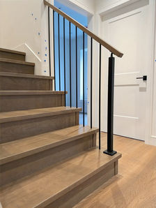 Modern interior staircase with light oak treads and risers, slim black metal balusters, square black newel post, and matching wood handrail beside a white shaker door.