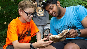 Two people outdoors examining a large mushroom. One holds a magnifying glass. Both wear shirts with tree patterns. Green foliage in background.