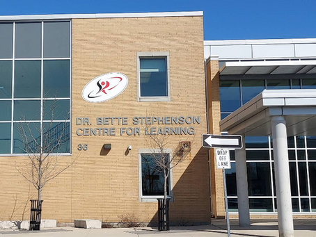 Modern brick building with large windows, reading "Dr. Bette Stephenson Centre for Learning." Clear blue sky, trees without leaves.