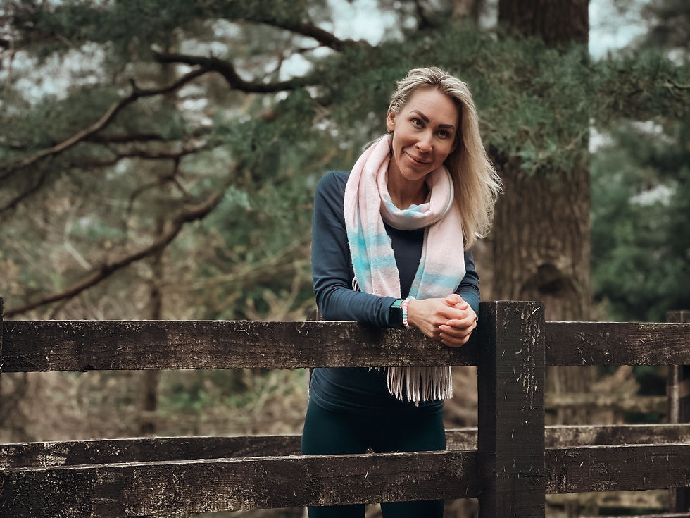 A woman stands on an old, tiny wooden bridge in a forest, looking away and smiling warmly. The scene is set on a beautiful autumn day in the Lake District, surrounded by vibrant fall foliage and a peaceful, natural atmosphere.