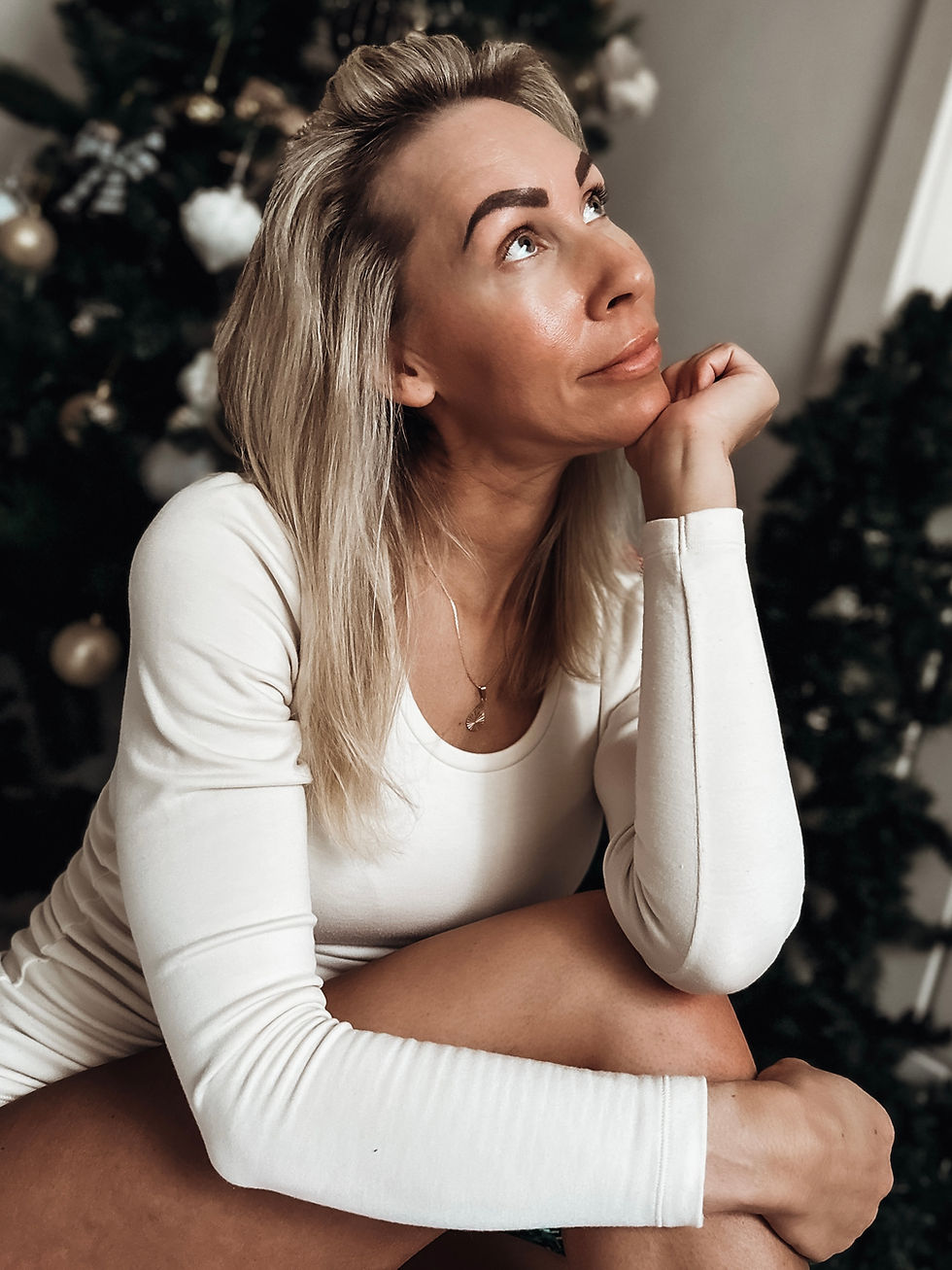 Lady sits next to Christmas tree looking thoughtful and smiles warmly
