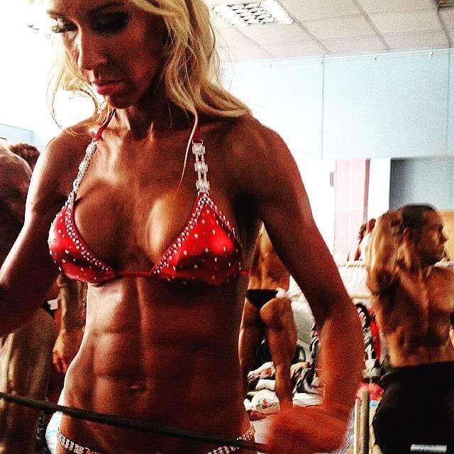 Woman stretching with an elastic band backstage at a bodybuilding, fitness, and bikini competition, competitors visible behind her, focused and preparing mindfully for the stage.