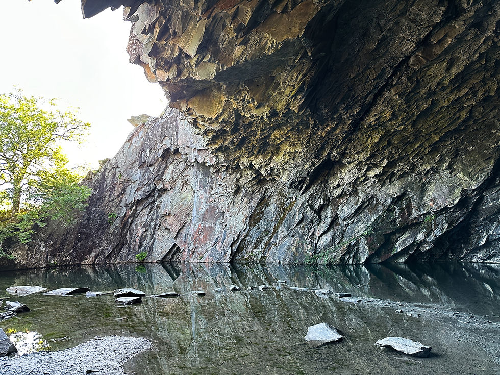 Inside Rydal Cave, Ambleside, Lake District, with sunlight streaming in and a line of stepping stones crossing the still cave water, leading towards the bright mountain view outside - a scene of calm and mindful presence.