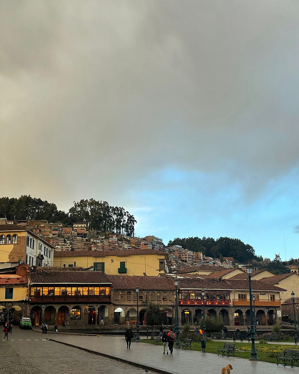 Historic plaza with arched buildings and hillside homes under a cloudy sky, people walking and a dog in the foreground
