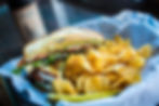 Grilled sandwich with lettuce and tomato next to a pile of potato chips on white paper. A pickle slice and blurry drink bottle in the background.